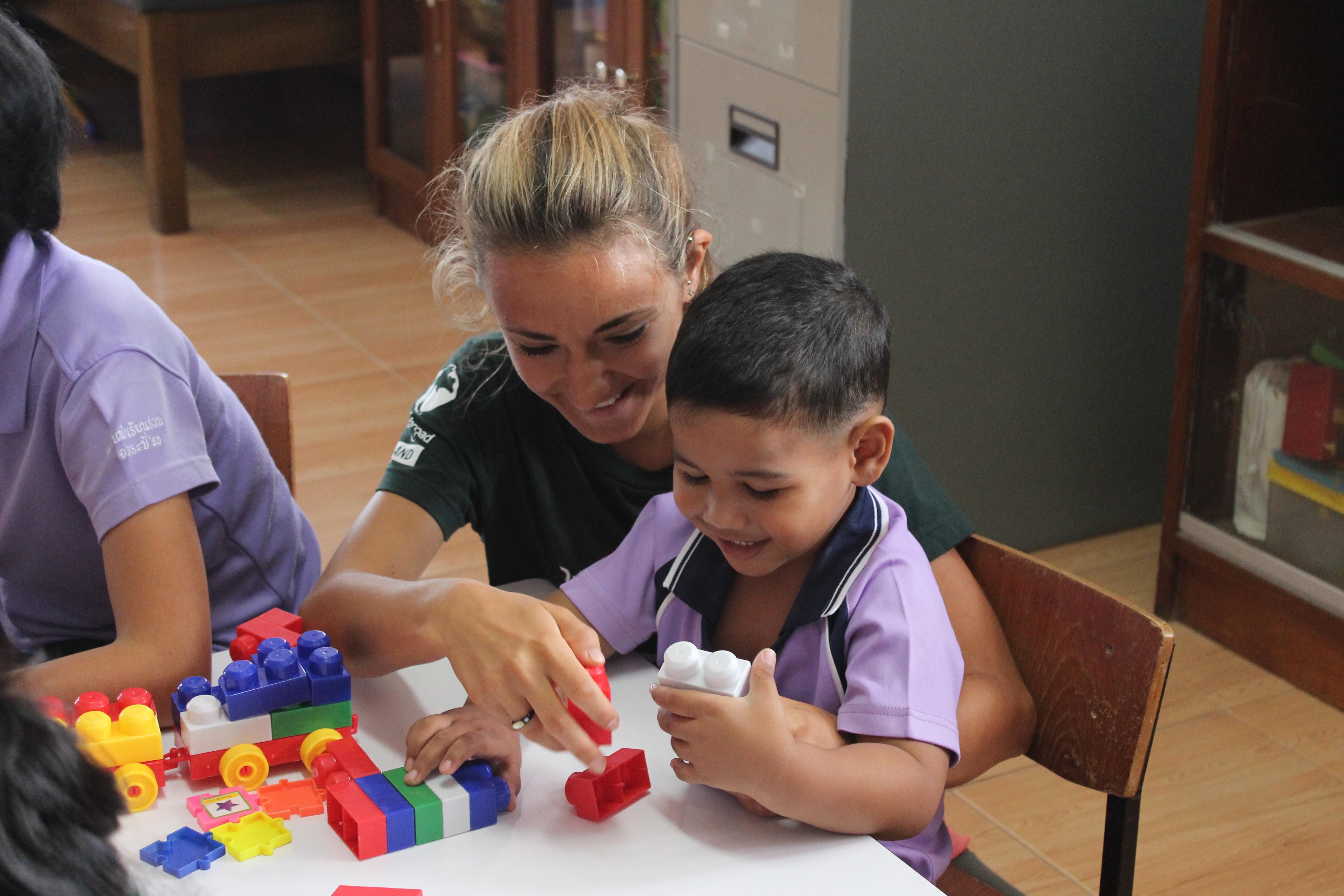 Projects Abroad volunteer working with children in Thailand plays lego with young boy during a care placement. Female Childcare volunteer helps young boy build lego blocks in a day care centre during Care placement in Thailand.